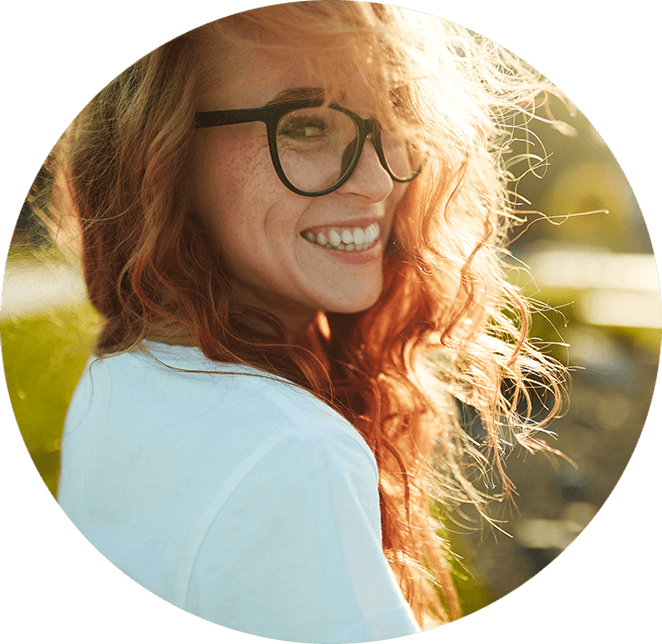 Very nice lady with red hair wearing glasses and white shirt smiling outside in a park with the sun shining on her